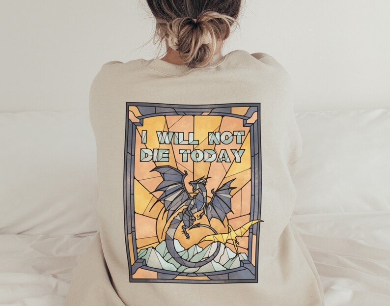 Pu&ograve; includere: Una felpa beige con un designo a vetri colorati che presenta un drago blu e il testo "I WILL NOT DIE TODAY".