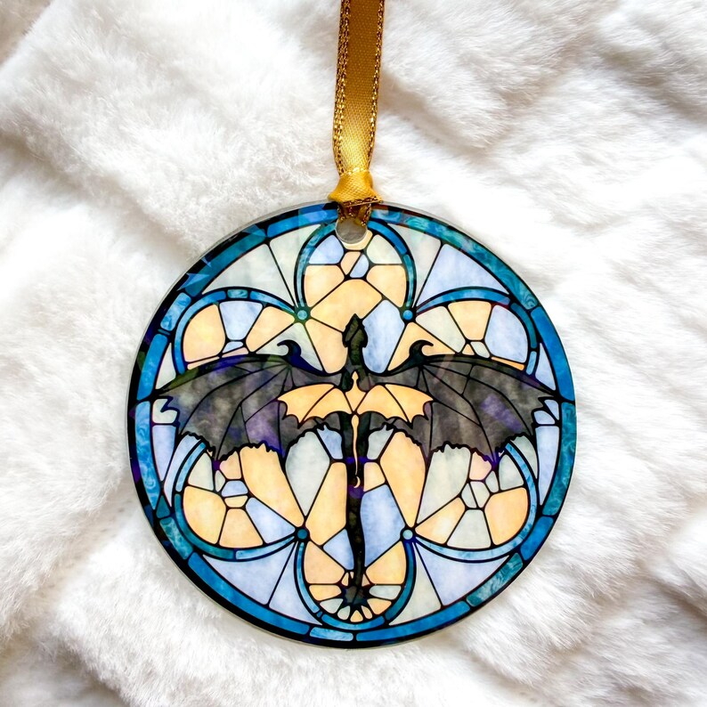 Op de afbeelding: Ronde ornament met een glas-in-lood ontwerp met een draak met uitgespreide vleugels. De draak is zwart en geel, tegen een achtergrond van blauwe, gele en witte geometrische vormen. Een gouden lint is bevestigd om op te hangen.
