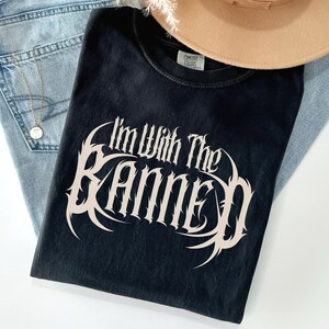 May include: Black t-shirt with the text "I'm With The Banned" in a stylized, off-white font. The shirt is folded and placed on top of a pair of blue jeans, a gold necklace, and a tan hat.
