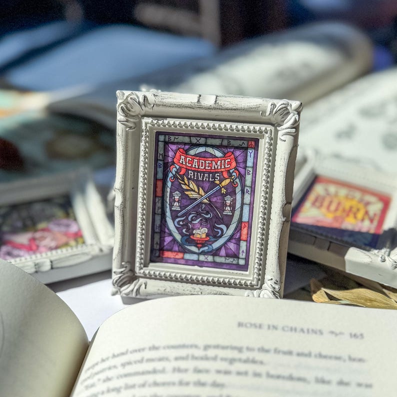 May include: A small, white, ornate picture frame with a stained-glass style image. The image features the words "Academic Rivals" above a quill and sword design. The frame is resting on a book with the title "Rose in Chains".