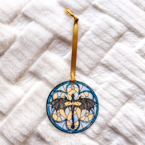 Op de afbeelding: Een ronde ornament met een glas-in-lood ontwerp met een draak met uitgespreide vleugels. Het ornament heeft een blauw, geel en zwart kleurenschema, met een gouden lint om op te hangen. De achtergrond is een zacht, wit, getextureerd oppervlak.