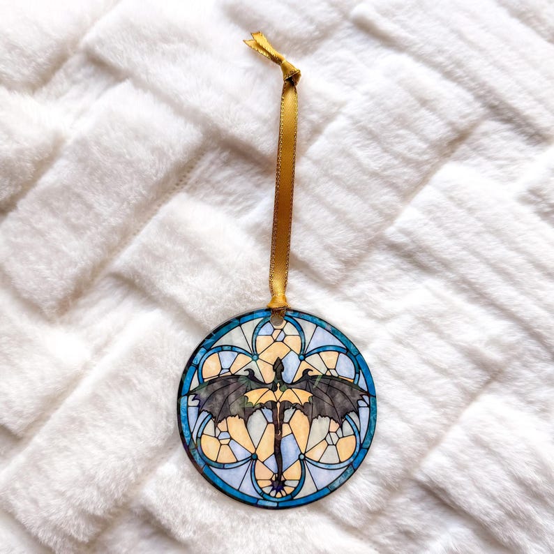 Op de afbeelding: Een ronde ornament met een glas-in-lood ontwerp met een draak met uitgespreide vleugels. Het ornament heeft een blauw, geel en zwart kleurenschema, met een gouden lint om op te hangen. De achtergrond is een zacht, wit, getextureerd oppervlak.
