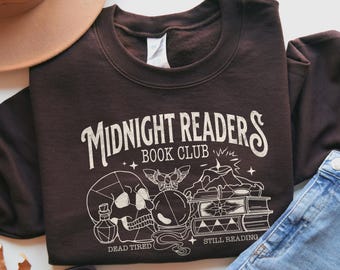 Puede incluir: Una sudadera marrón oscuro con la frase "Midnight Readers Book Club" en una fuente vintage. El diseño incluye una calavera, libros, una bola de cristal y los textos "Dead Tired" y "Still Reading".