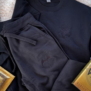 K&ouml;nnte beinhalten: Schwarze Jogginghose und ein schwarzer Pullover mit einer Grafik eines Vogels und dem Text "For The Glory Of The Underworld".