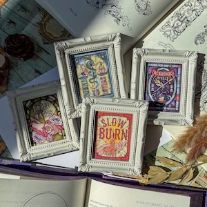 May include: Four small, white framed art pieces with ornate edges. Each frame contains a colorful, stained-glass style design with text: "Fated Mates," "Lovers," "Slow Burn," and "Academic Rivals." The art pieces are arranged on a table with an open book.