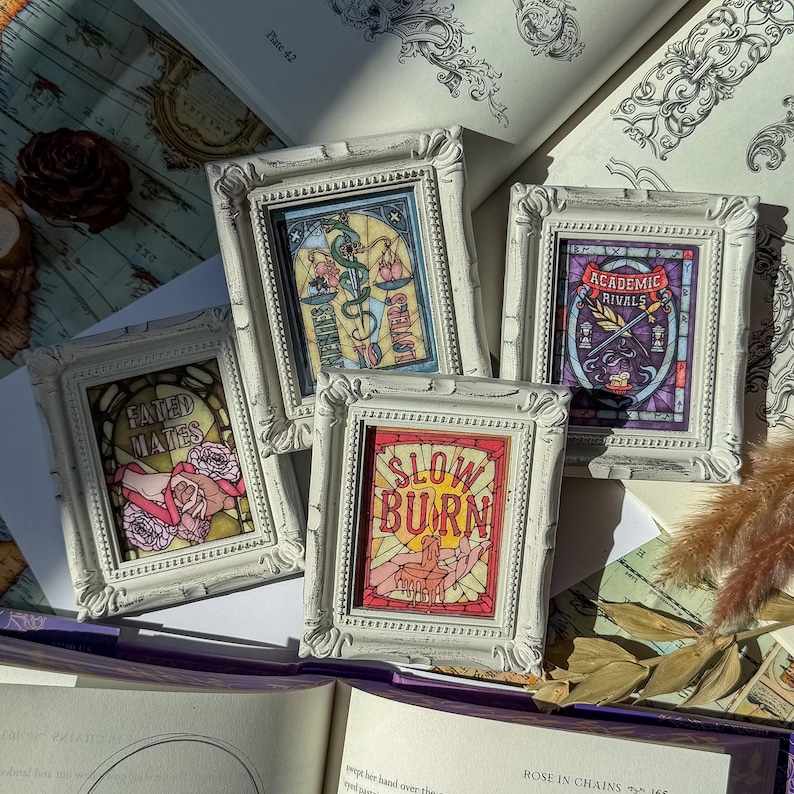 May include: Four small, white framed art pieces with ornate edges. Each frame contains a colorful, stained-glass style design with text: "Fated Mates," "Lovers," "Slow Burn," and "Academic Rivals." The art pieces are arranged on a table with an open book.