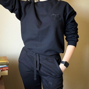 K&ouml;nnte beinhalten: Schwarzer Pullover und Jogginghose mit einem schwarzen gestickten Design eines Vogels mit dem Text "Theology of the Birds" auf der Vorder- und R&uuml;ckseite.