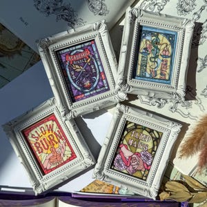 May include: Four small, white framed art pieces with stained glass style designs. Each frame features a different design with text: "Academic Rivals", "Enemies Lovers", "Slow Burn", and "Fated Mates". The art pieces are in a vintage style.
