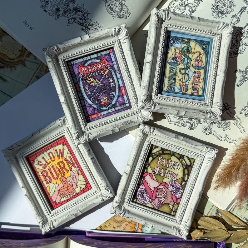 May include: Four small, white framed art pieces with stained glass style designs. Each frame features a different design with text: "Academic Rivals", "Enemies Lovers", "Slow Burn", and "Fated Mates". The art pieces are in a vintage style.