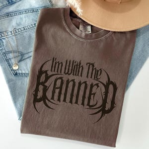 May include: A brown t-shirt with the text "I'm With The Banned" in a gothic font. The shirt is folded and placed on top of a pair of blue jeans, with a gold necklace and a tan hat visible.