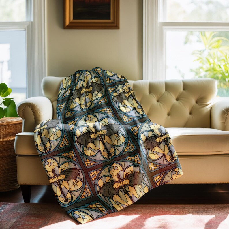 May include: A decorative throw blanket featuring a repeating pattern of bats in a stained-glass style. The design incorporates shades of black, gray, yellow, and blue, creating a gothic aesthetic. The blanket is draped over a cream-colored armchair.