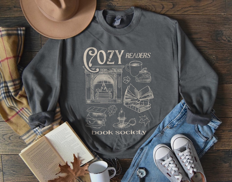 May include: A gray sweatshirt with the text 'Cozy Readers Book Society' printed on the front. The design includes a fireplace, books, a cup of coffee, and a candle. The sweatshirt is laid out on a wooden surface with a plaid blanket, a book, a hat, and jeans.