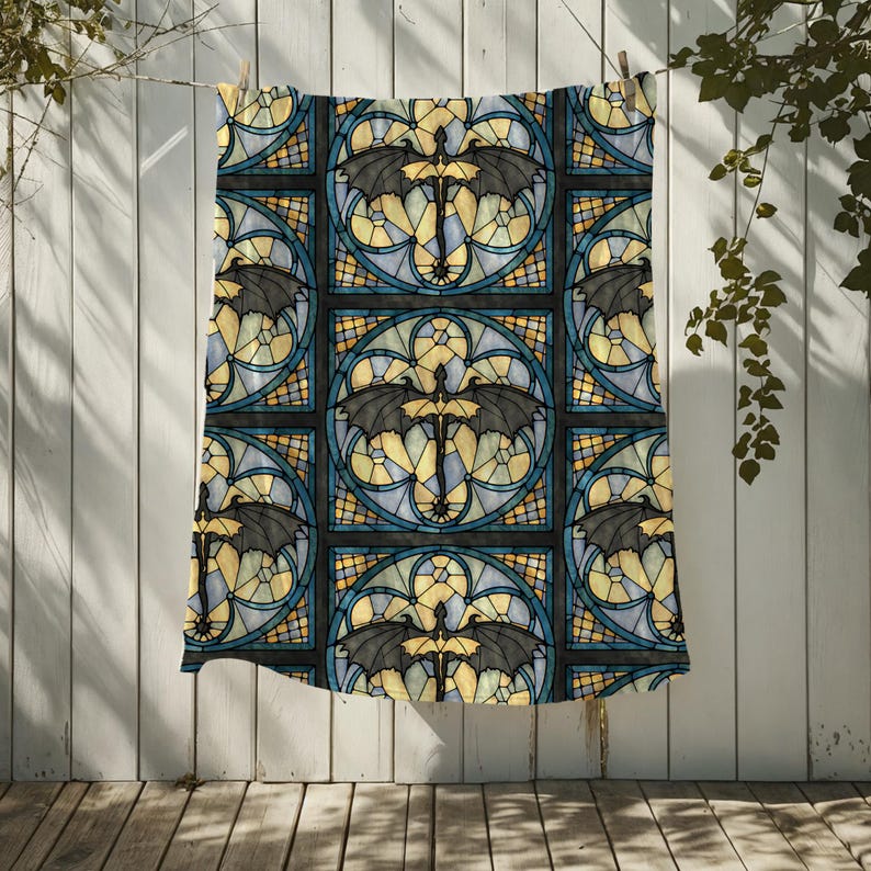 May include: A patterned textile featuring a stained-glass design with black bat-like creatures. The design incorporates yellow, blue, and black colors, creating a gothic or fantasy theme. The textile is hanging on a clothesline against a white wooden background.