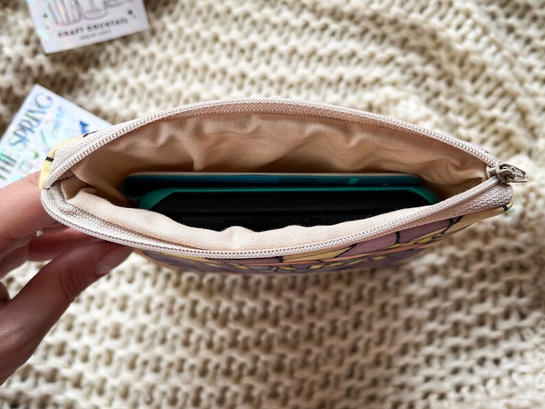 May include: A small zippered pouch with a floral print and a light brown lining. A teal and black electronic device is inside the pouch.