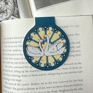 May include: A teal bookmark featuring a stained-glass design with two white swans facing each other. The swans have red beaks and are set against a yellow and blue background. The bookmark is placed in an open book.