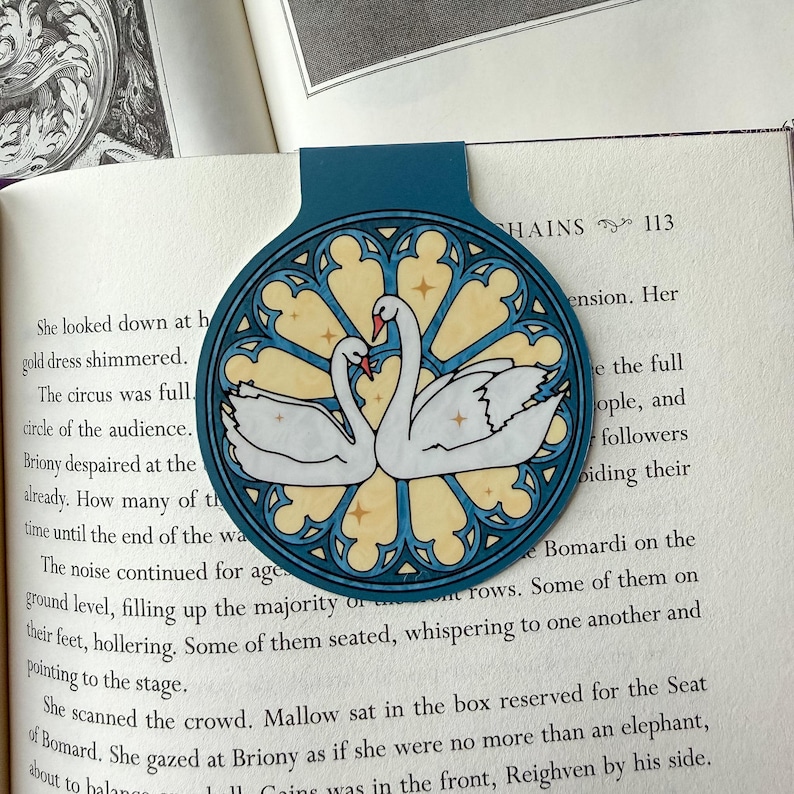 May include: A teal bookmark featuring a stained-glass design with two white swans facing each other. The swans have red beaks and are set against a yellow and blue background. The bookmark is placed in an open book.