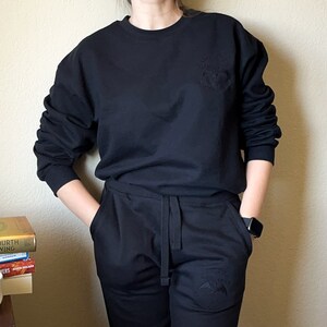 K&ouml;nnte beinhalten: Ein schwarzer Pullover mit einem kleinen gestickten Design auf der Vorderseite und passende schwarze Jogginghose mit einem &auml;hnlichen Design am linken Bein.