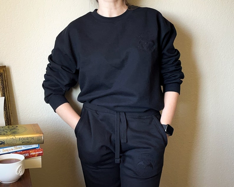 K&ouml;nnte beinhalten: Ein schwarzer Pullover mit einem kleinen gestickten Design auf der Vorderseite und passende schwarze Jogginghose mit einem &auml;hnlichen Design am linken Bein.