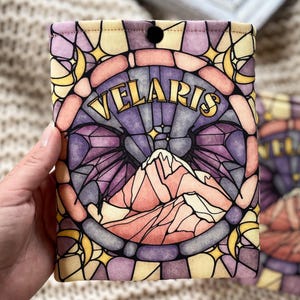 May include: A yellow, purple, and pink stained glass pattern book sleeve with the word "VELARIS" in gold lettering. The design features a mountain range and crescent moons.