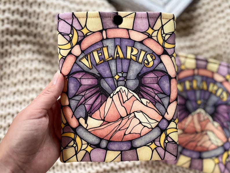May include: A yellow, purple, and pink stained glass pattern book sleeve with the word "VELARIS" in gold lettering. The design features a mountain range and crescent moons.