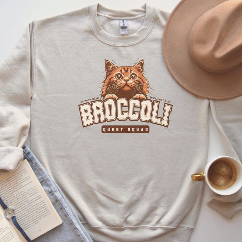 Pu&ograve; includere: Una felpa beige chiaro con una grafica marrone di un gatto che sporge sopra il testo "BROCCOLI QUEST SQUAD". Il testo "no one wants it" &egrave; a sinistra della grafica e il testo "but it's good for you" &egrave; a destra della grafica.