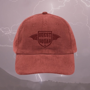 以下が含まれることがあります： 赤いコーデュロイのベースボールキャップ。黒い刺繍で「QUEST SQUAD」とあり、盾の両側に翼がついている。