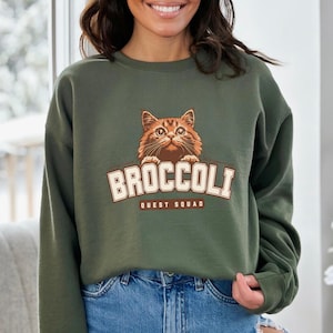 Pu&ograve; includere: Una felpa verde con una grafica di un gatto che sbircia da sopra con il testo "Broccoli Quest Squad" stampato sul davanti.