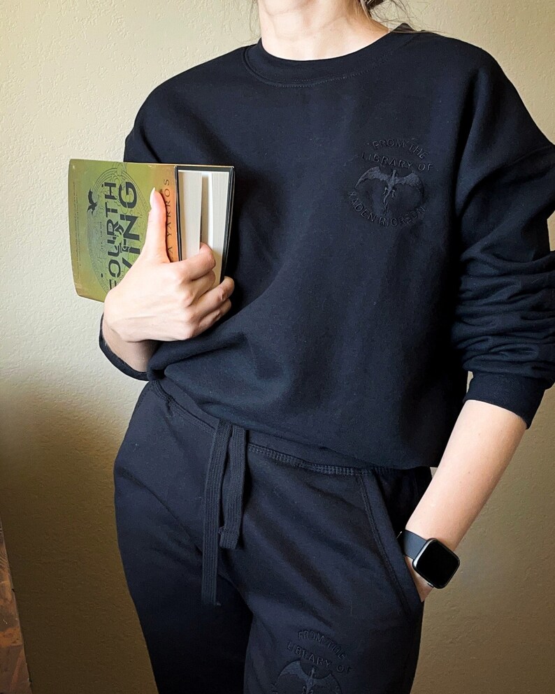 K&ouml;nnte beinhalten: Eine Person tr&auml;gt ein schwarzes Sweatshirt mit einem schwarzen Fledermaus-Motiv und dem Text "From the Library of Shadows" auf der Vorderseite. Die Person tr&auml;gt auch eine schwarze Jogginghose mit einem &auml;hnlichen Fledermaus-Motiv und Text auf dem linken Bein. Die Person h&auml;lt ein Buch mit einem gelben und gr&uuml;nen Einband, auf dem "Fourth Wing" steht.