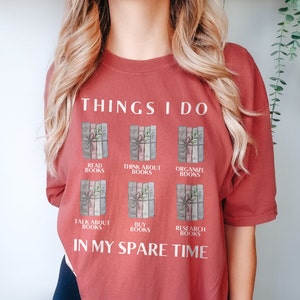 Może przedstawiać: Koszulka w kolorze rdzy z tekstem "THINGS I DO" i grafiką sześciu stos&oacute;w książek z tekstem opisującym czynności związane z książkami, takie jak "READ BOOKS", "THINK ABOUT BOOKS", "ORGANIZE BOOKS", "TALK ABOUT BOOKS", "BUY BOOKS" i "RESEARCH BOOKS". Tekst "IN MY SPARE TIME" znajduje się pod grafiką.