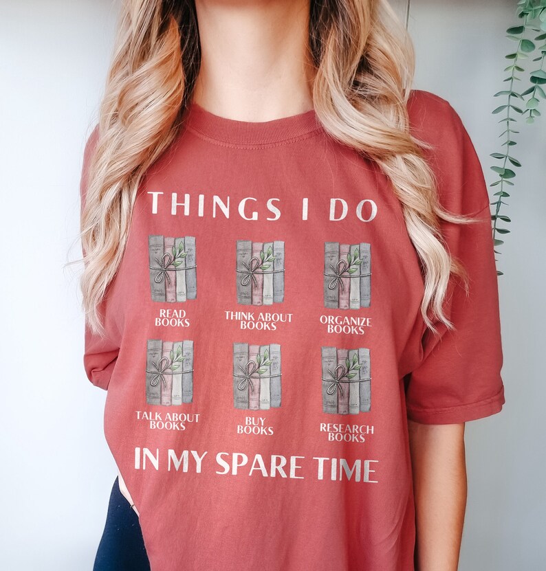 Może przedstawiać: Koszulka w kolorze rdzy z tekstem "THINGS I DO" i grafiką sześciu stos&oacute;w książek z tekstem opisującym czynności związane z książkami, takie jak "READ BOOKS", "THINK ABOUT BOOKS", "ORGANIZE BOOKS", "TALK ABOUT BOOKS", "BUY BOOKS" i "RESEARCH BOOKS". Tekst "IN MY SPARE TIME" znajduje się pod grafiką.