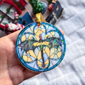 Op de afbeelding: Ronde ornament met een glas-in-lood ontwerp met een draak. Het ornament heeft een blauwe rand en een gouden lint om op te hangen. De draak is afgebeeld met zwarte vleugels en een gele romp, tegen een achtergrond van gele en blauwe geometrische vormen.