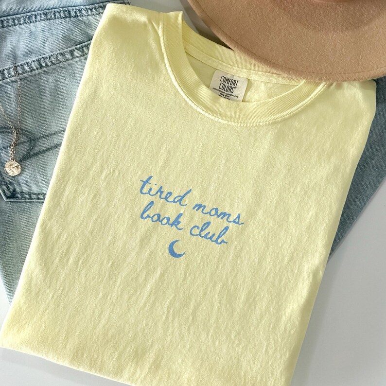 May include: A light yellow t-shirt with the text "tired moms book club" in blue script. The shirt is folded on top of blue jeans and a tan hat. The shirt has a Comfort Colors tag.