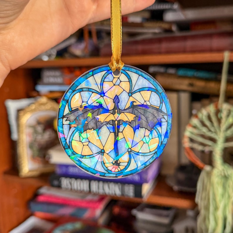 Op de afbeelding: Een rond ornament in glas-in-loodstijl met een drakenontwerp. Het heeft een gouden lint om op te hangen. De draak heeft zwarte vleugels tegen een achtergrond van geel, blauw en iriserende accenten. De diameter is ongeveer 8 cm.