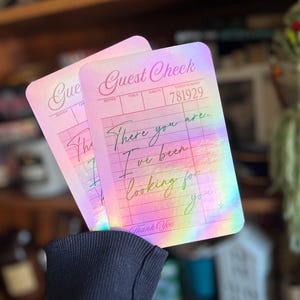 May include: A holographic pink and blue guest check with the text "Guest Check" and "There you are. I've been looking for you." written in black cursive.