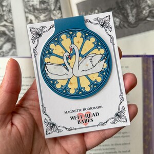 May include: A magnetic bookmark featuring a stained-glass design with two white swans. The bookmark has a teal top and is on a white card with the text "MAGNETIC BOOKMARK WELL READ BABES". The design is set against a yellow background.