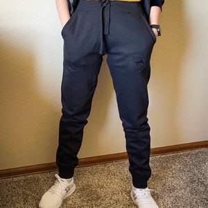 K&ouml;nnte beinhalten: Schwarze Jogginghose mit Kordelzugbund und kleinem gesticktem Logo am rechten Bein. Die Jogginghose ist an den Kn&ouml;cheln umgeschlagen.