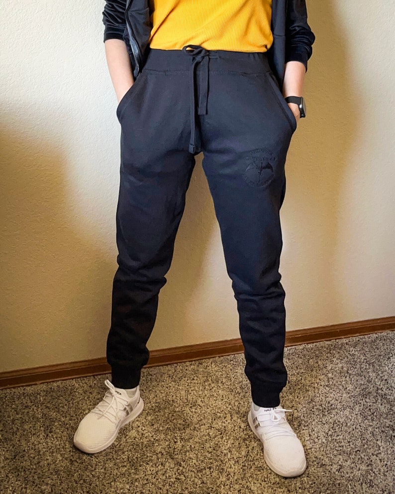 K&ouml;nnte beinhalten: Schwarze Jogginghose mit Kordelzugbund und kleinem gesticktem Logo am rechten Bein. Die Jogginghose ist an den Kn&ouml;cheln umgeschlagen.