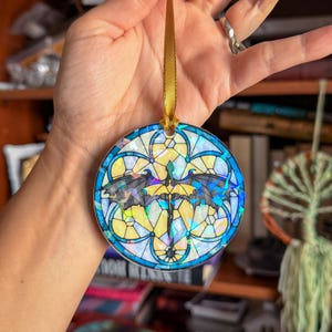 Op de afbeelding: Ronde ornament met een glas-in-lood ontwerp met een draak. Het ornament heeft een blauwe rand en een gouden lint om op te hangen. De draak is zwart en geel, met gedetailleerde vleugels. De achtergrond is een mix van geel, wit en blauw.
