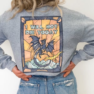 Pu&ograve; includere: Una felpa grigia con un design a vetri colorati che presenta un drago e il testo "I WILL NOT DIE TODAY".