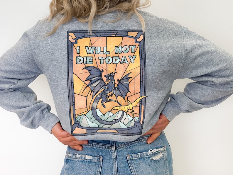 Pu&ograve; includere: Una felpa grigia con un design a vetri colorati che presenta un drago e il testo "I WILL NOT DIE TODAY".