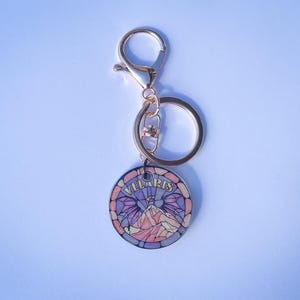 Op de afbeelding: Een gouden sleutelhanger met een ronde hanger met een glas-in-loodontwerp van een bergketen met vleugels en het woord "VELLARIS" in gouden letters.