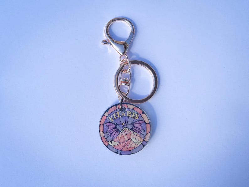 Op de afbeelding: Een gouden sleutelhanger met een ronde hanger met een glas-in-loodontwerp van een bergketen met vleugels en het woord "VELLARIS" in gouden letters.
