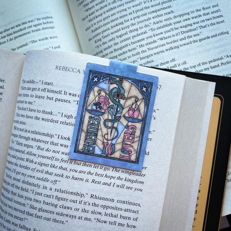 K&ouml;nnte beinhalten: Ein blaues und pinkfarbenes Lesezeichen mit einem Buntglasdesign. Das Lesezeichen zeigt eine Waage mit einem Schwert und die Worte "Enemies to Lovers".