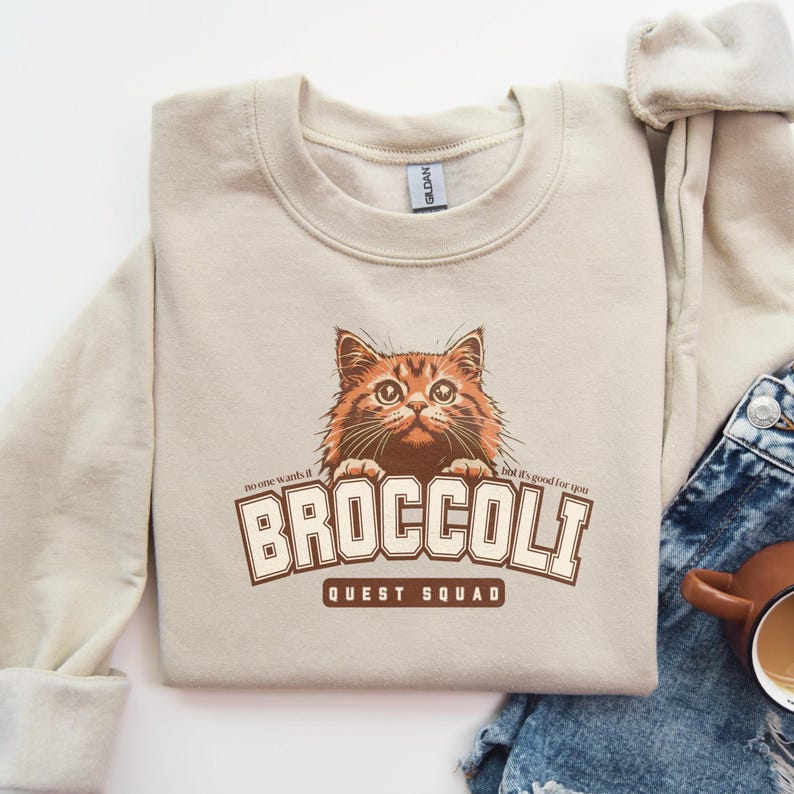 Pu&ograve; includere: Una felpa beige con una grafica di un gatto che spunta da sopra il testo "BROCCOLI QUEST SQUAD". Il testo "no one wants it" &egrave; sopra il gatto e il testo "but it's good for you" &egrave; sotto il gatto.