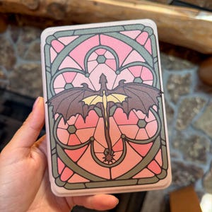 Könnte beinhalten: Ein rechteckiges Etui mit einem Buntglasfenster-Design in Rosa- und Orangetönen. Ein dunkelbrauner Drache mit ausgebreiteten Flügeln und einem blassgelben Körper ist mittig im Rahmen platziert, umgeben von dunkelgrauen Bleilinien.