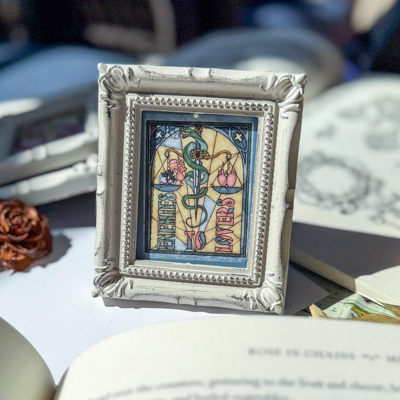 May include: A small, ornate, white-framed artwork featuring a stained-glass style design. The artwork depicts a caduceus symbol with scales and the words "Enemies to Lovers". The frame has a distressed, vintage appearance.