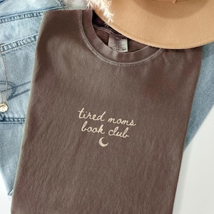 May include: A brown t-shirt with the text "tired moms book club" in white script. The shirt is folded and rests on a pair of blue jeans. A tan hat is visible in the background.