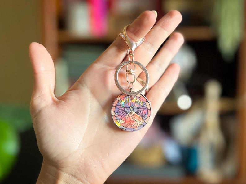 Op de afbeelding: Een zilveren sleutelhanger met een ronde, kleurrijke hanger met de tekst "Wild & Free". De hanger heeft een glas-in-loodontwerp met een roze, paarse en blauwe kleurstelling.