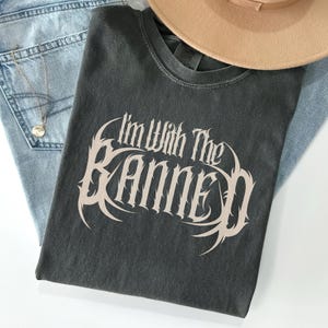 May include: Dark gray t-shirt with the text "I'm With The Banned" in a stylized, off-white font. The shirt is laid on top of light blue jeans and a tan hat. A silver necklace with a pendant is visible.
