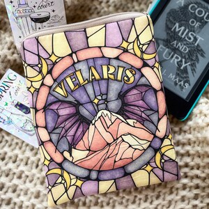 May include: A stained glass style book sleeve with a purple, pink, and yellow design. The sleeve features a mountain range and the words "VELARIS" in gold lettering.